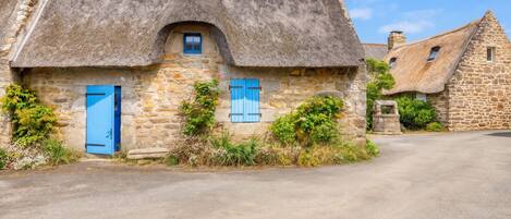 A stone cottage with a thatched roof, blue shutters, and a small window.