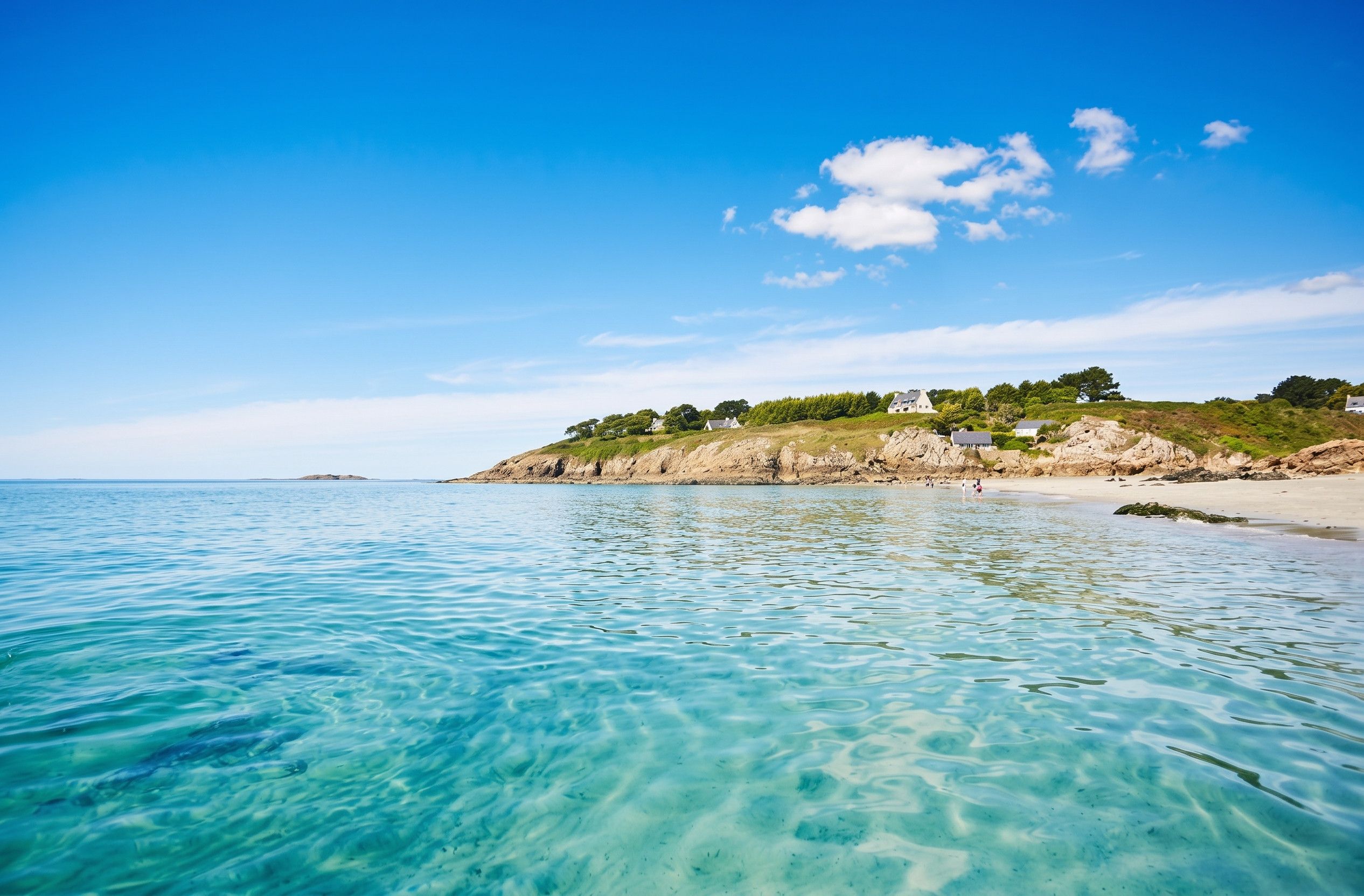 A coastal scene with clear turquoise water, a sandy beach, and a rocky hillside with buildings.