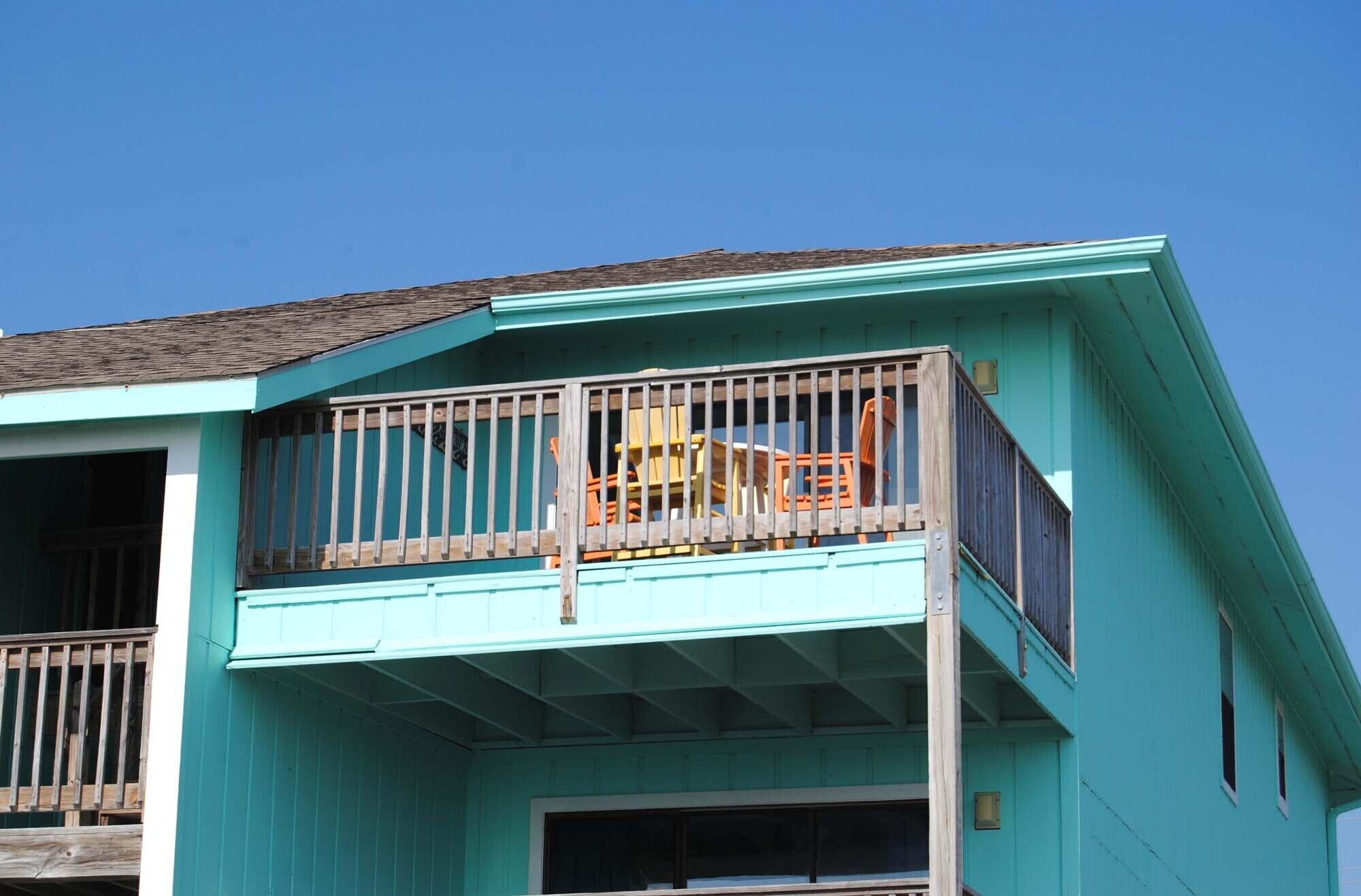 A turquoise two-story building with a balcony featuring a wooden railing and a yellow chair.