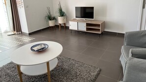Una sala de estar moderna con una televisión de pantalla plana sobre un mueble de madera, una mesa redonda blanca con un control remoto, dos plantas en macetas y un sofá gris.