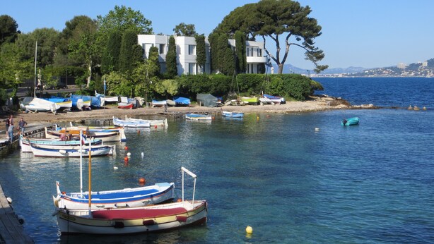 Una zona costera con barcos amarrados a la orilla, un edificio con balcón con vistas al agua, y una vista despejada del mar.
