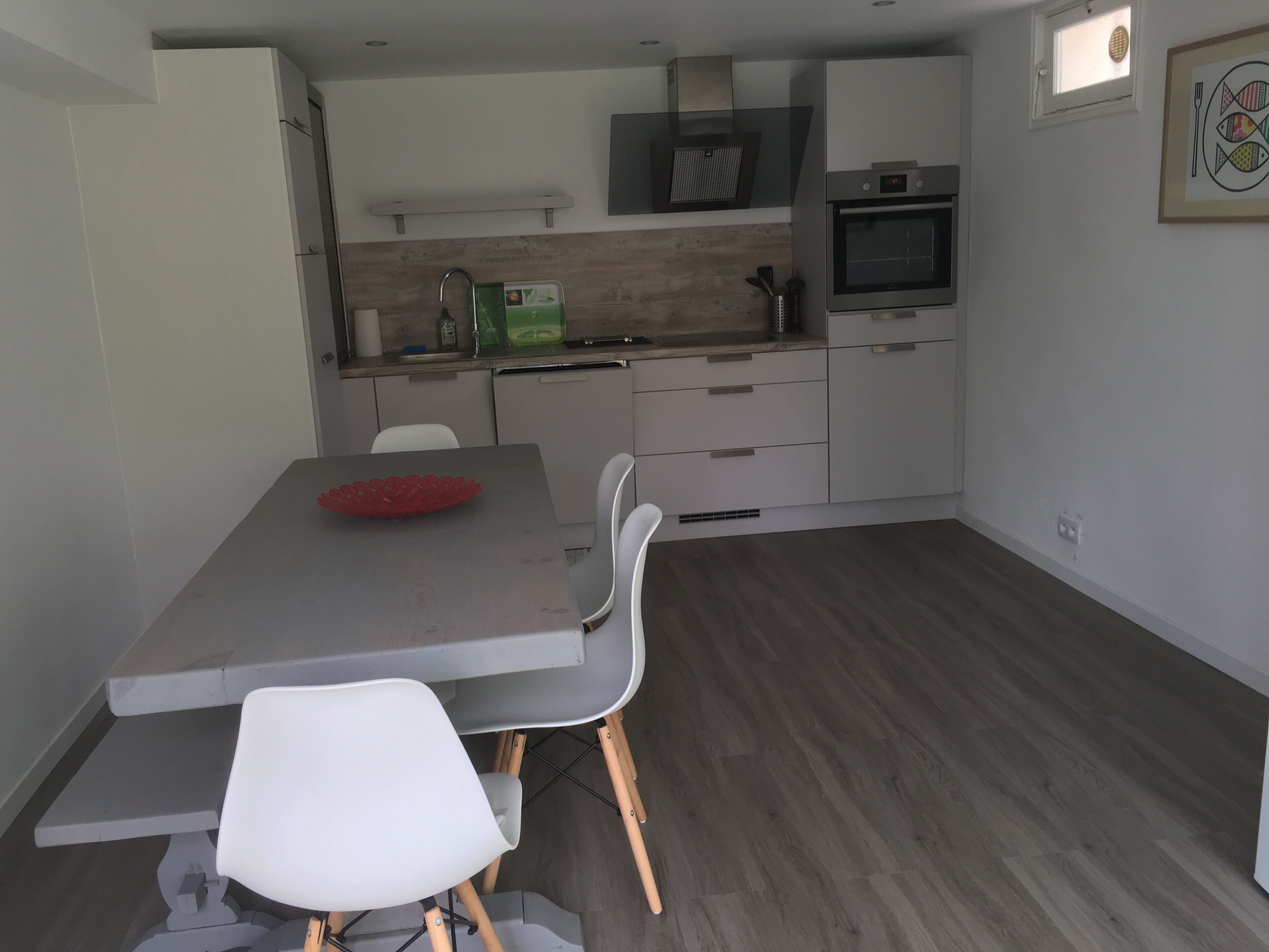 Une cuisine moderne avec des meubles blancs, un four encastré et un coin repas comprenant une table grise et des chaises blanches.