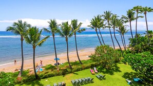 A beach view with palm trees, a grassy area with lounge chairs, and a sandy beach. - Stunning Views & Tropical Tranquility 429 LS (Lahaina)