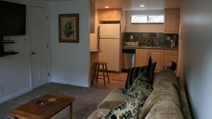 A compact living space with a sofa, a small table, a kitchen area with cabinets and a refrigerator, and a television mounted on the wall. - Steps from watching Sunsets in the Sand (Carpinteria)