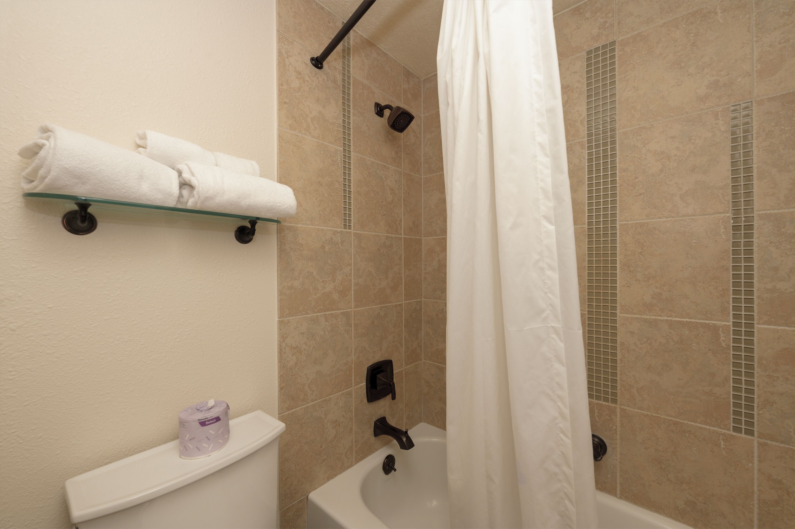 A bathroom with a glass shelf holding towels, a toilet paper holder, and a bathtub with a shower curtain.