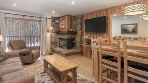 A cozy living room with a stone fireplace, a comfortable sofa, a wooden dining table with chairs, and a television mounted on the wall. - Beaver Village Unit #1612 Value priced 2 bed great for families & quick getaways (Winter Park)