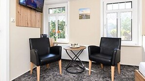 Et værelse med to sorte læderstole, et lille rundt bord, et fjernsyn monteret på væggen og et vindue med udsigt til grønne områder.