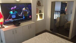Ein Fernsehgerät zeigt ein Sportevent, ein Bett, ein Kleiderschrank und ein Spiegel, der einen Raum widerspiegelt.