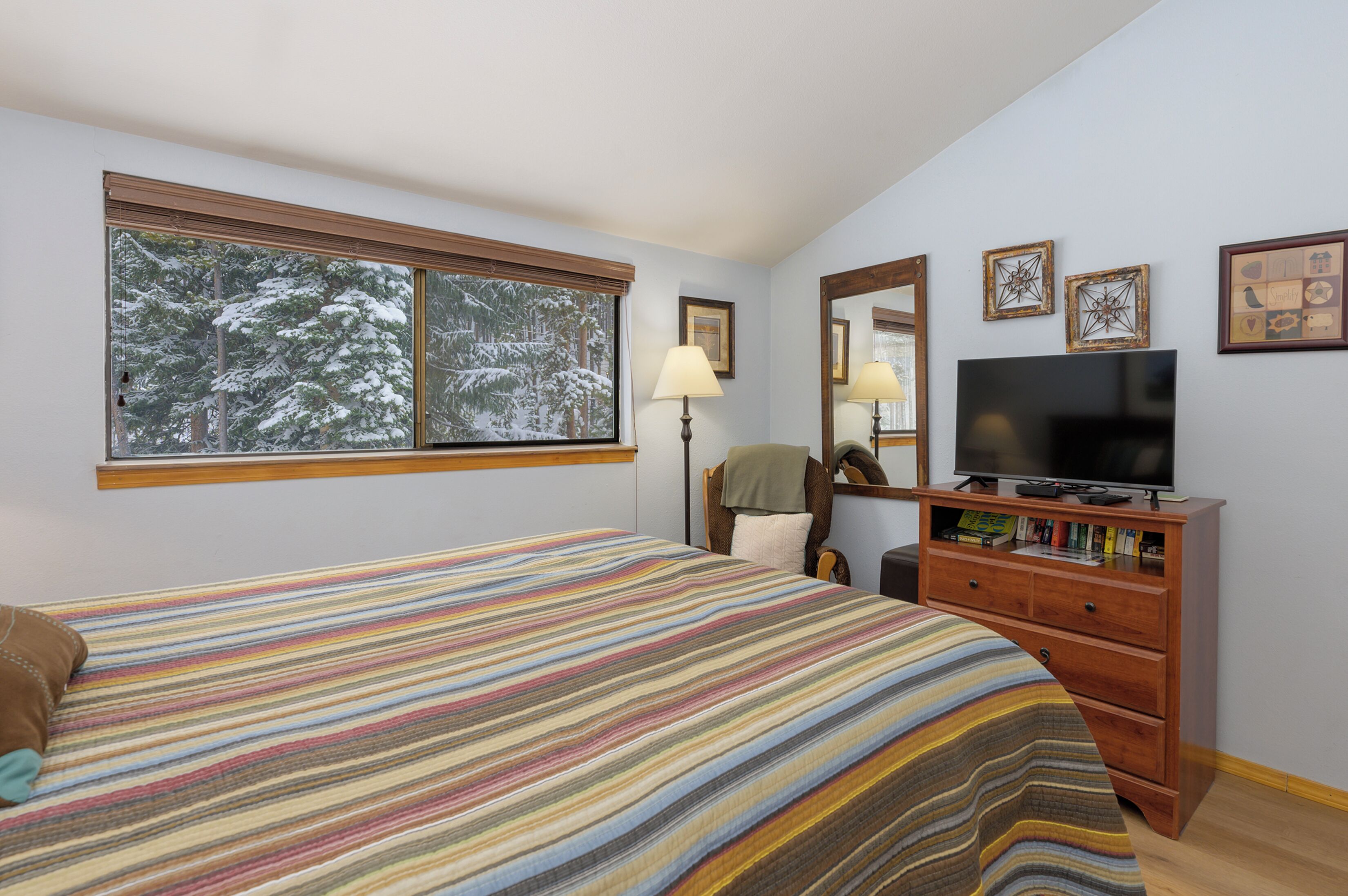 A bedroom with a large window, a bed with a striped blanket, a dresser with a TV, and framed pictures on the wall.
