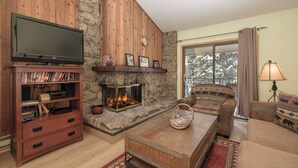 A cozy living room with a fireplace, a television on a wooden stand, a couch, and a window with a view of trees.