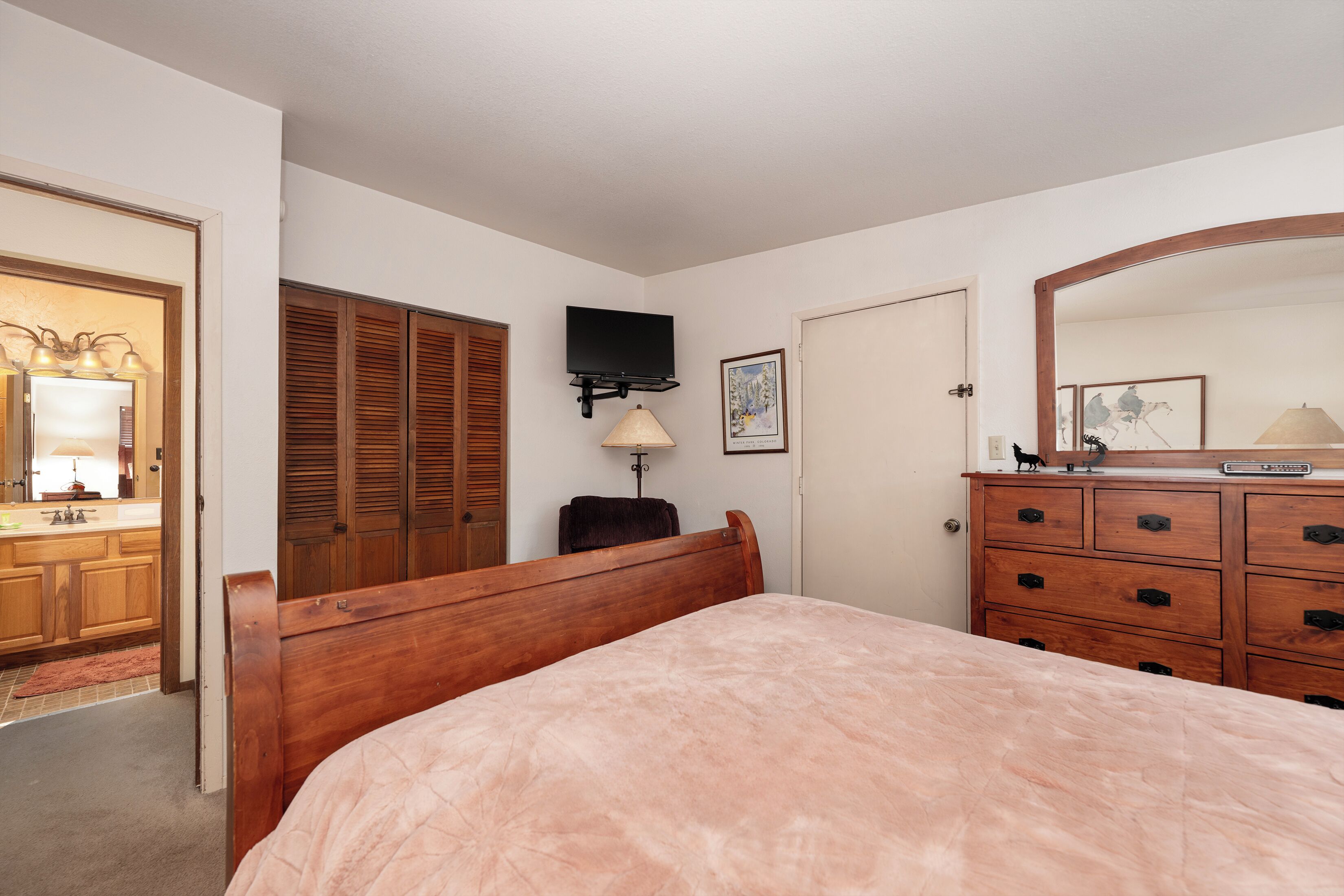 Een slaapkamer met een houten bed, een dressoir, een televisie en een deur met een spiegel.