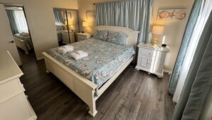 A bedroom with a bed, a nightstand, a ceiling fan, a mirror, and a window with curtains. - Spacious, Updated Villa – Perfect for Family Beach Vacations, Two King Beds! (Hilton Head Island)