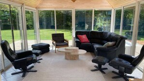 A sunroom with a wooden ceiling, large glass doors, a black leather sofa, and matching chairs.
