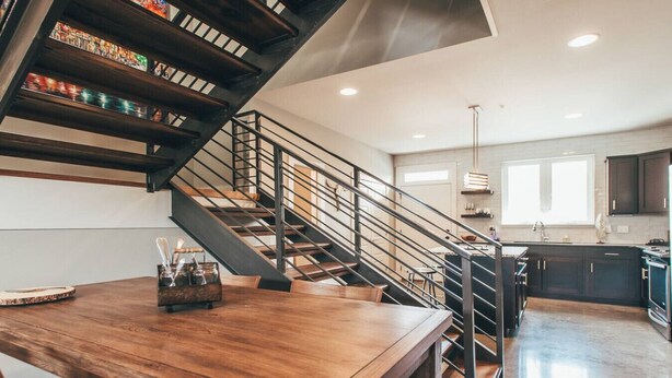 A modern kitchen with dark cabinets, a wooden dining table, and a staircase with metal railings. - Spacious Cowboy home near East Nashville with rooftop and views (Nashville)