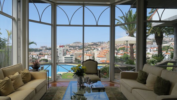 A spacious room with a large bay window offering a view of a cityscape and a pool. - villa in the center of Funchal with pool and garden (Funchal)