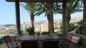 A covered outdoor seating area with a table and chairs, surrounded by palm trees, overlooking a cityscape with buildings and a view of the sea. - villa in the center of Funchal with pool and garden (Funchal)