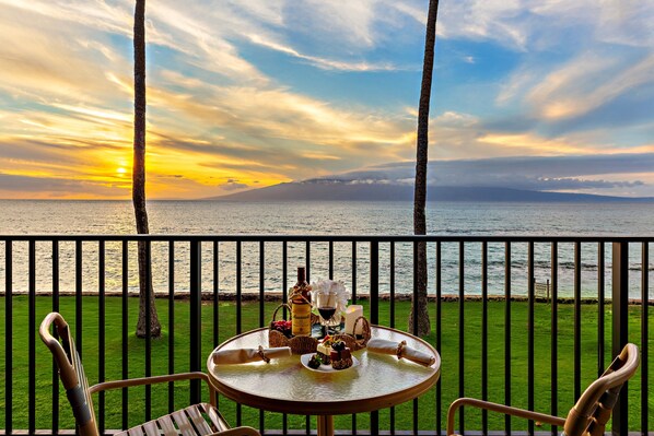 A table with a bottle, glasses, and a candle on a balcony overlooking a body of water with a sunset.