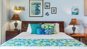 A neatly made bed with a tropical-themed bedspread, a wooden headboard, and a wall adorned with framed artwork and a decorative sign. - Luxury oceanfront studio with spacious lanai (Lahaina)