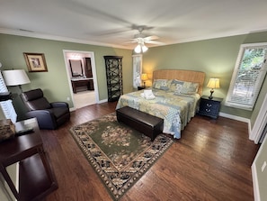 A bedroom with a bed, a chair, a desk, a nightstand, a lamp, a window with blinds, and a ceiling fan.