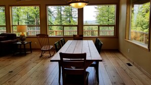 Zona pranzo con tavolo e sedie in legno, ampie finestre vista alberi e lampadario a sospensione.
