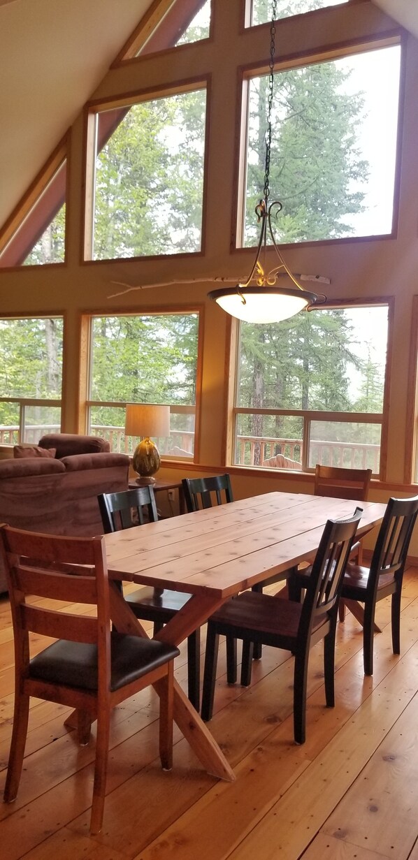 Un comedor con mesa y sillas de madera, ventanas amplias con vistas a árboles y una lámpara colgante.