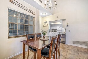 Un comedor con una mesa y sillas de madera, una ventana con persianas, y una cocina visible a través de un vano abierto.