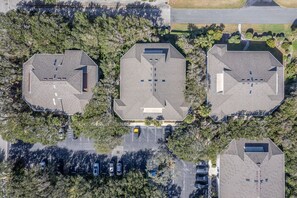 Vista aérea de un complejo de edificios con tres estructuras principales, cada una con techo plano y múltiples ventanas. Hay un estacionamiento frente a los edificios, y el área está rodeada de árboles.