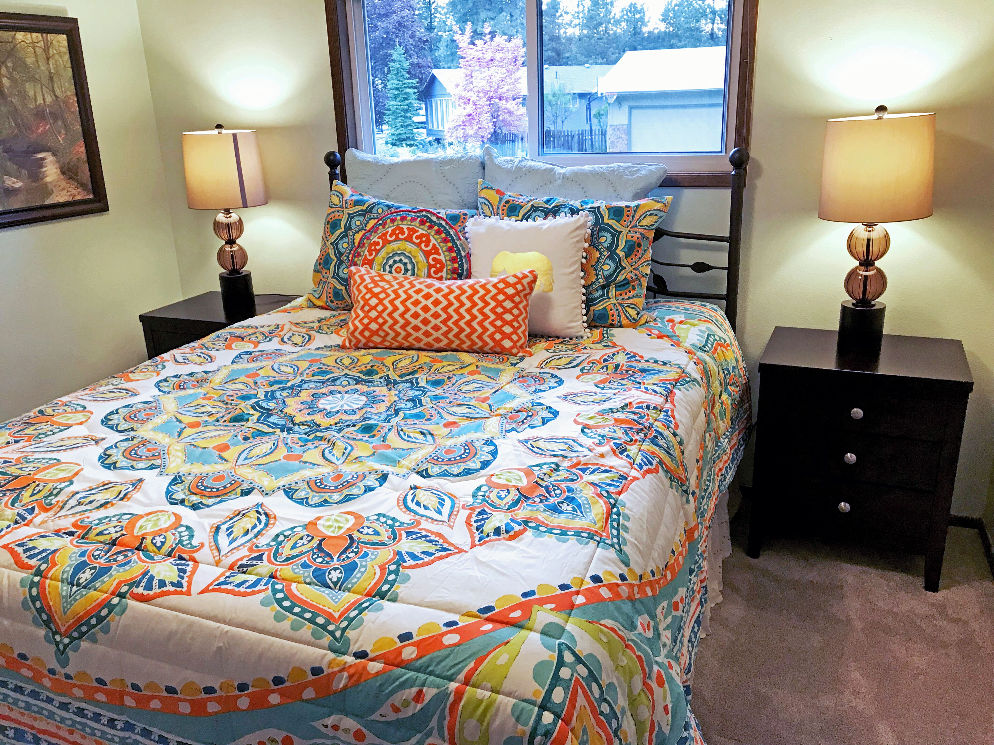 A neatly made bed with a colorful patterned bedspread, two bedside tables with lamps, and a window with a view of trees and a house.