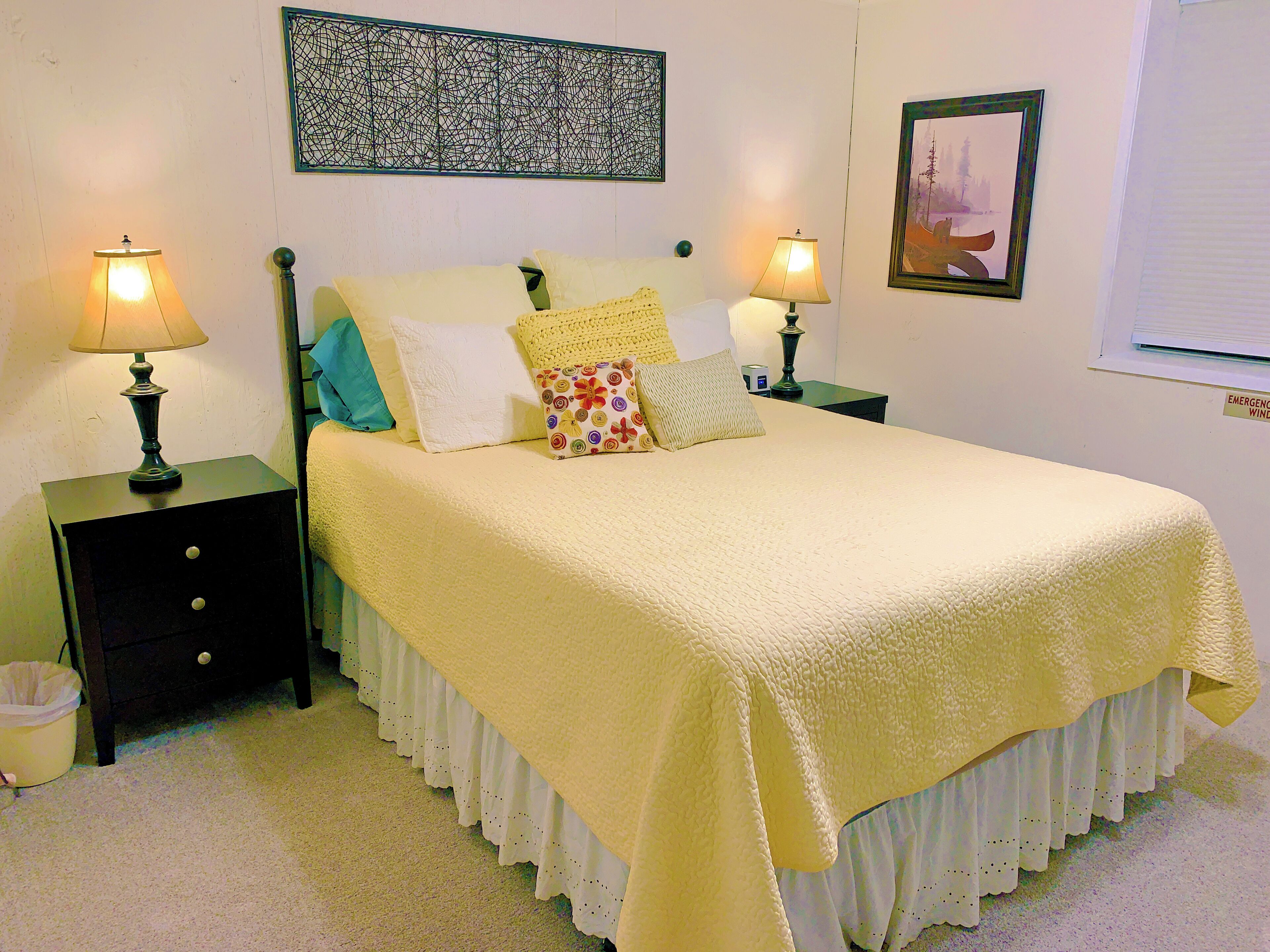 A neatly made bed with a yellow bedspread and multiple pillows, two bedside tables with lamps, a wall art piece, and a framed picture.