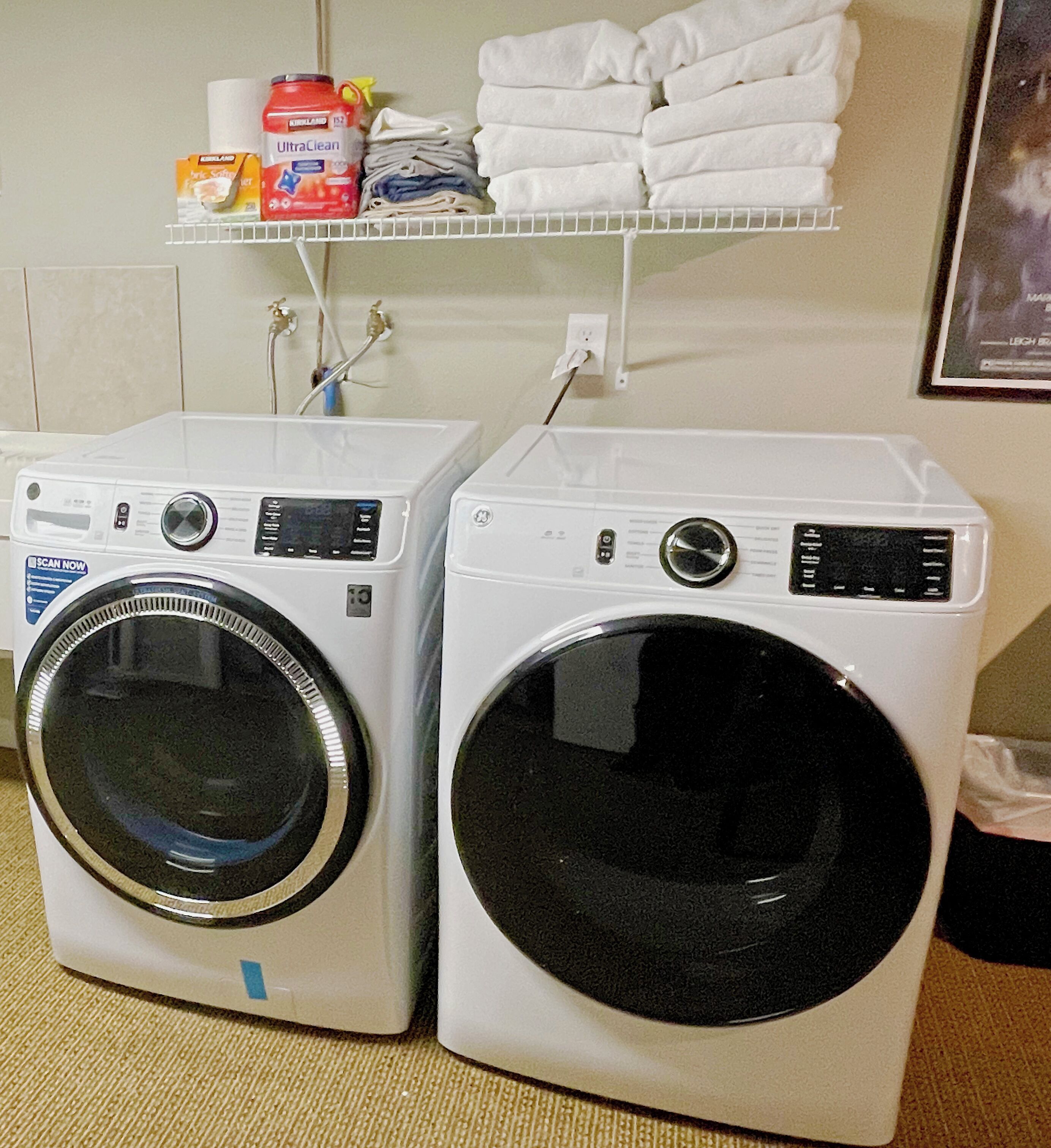 Two front-loading washing machines side by side, a shelf with folded towels and cleaning supplies, and a wall socket.