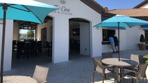 Ein Außenbereich mit Tischen und Stühlen unter blauen Sonnenschirmen, ein weißes Gebäude mit der Beschriftung "Fi Casa Bar & Lounge" und einem strahlend blauen Himmel.