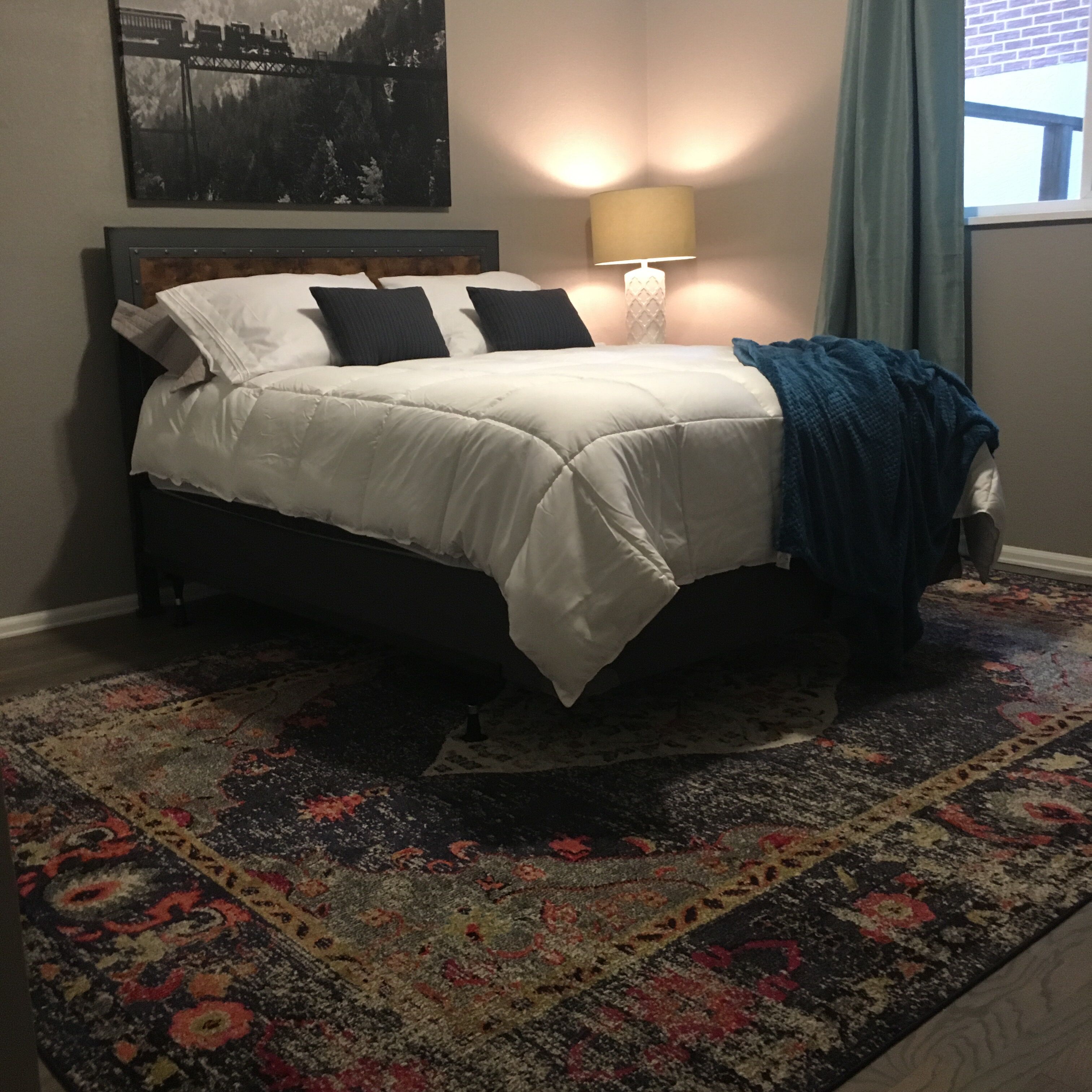 A bed with white bedding, a patterned rug, a lamp, and a painting on the wall.