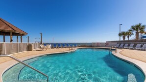 Una piscina azul cristalina con una terraza de madera, sillas de descanso y una zona de estar cubierta.