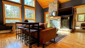 Una sala de estar acogedora con chimenea de piedra, pisos de madera y ventanas amplias con vistas a árboles.