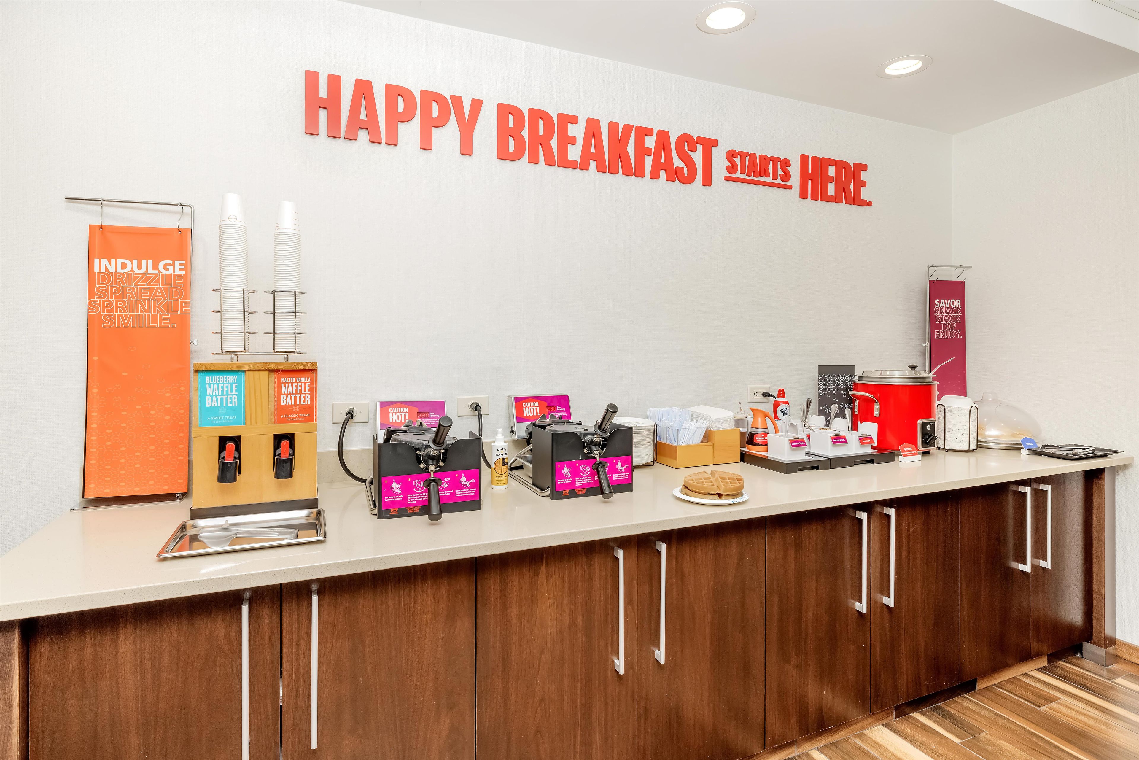 Una estación de desayuno con máquinas para hacer gofres, dispensadores de masa para gofres y un letrero que dice 'HAPPY BREAKFAST START HERE'.