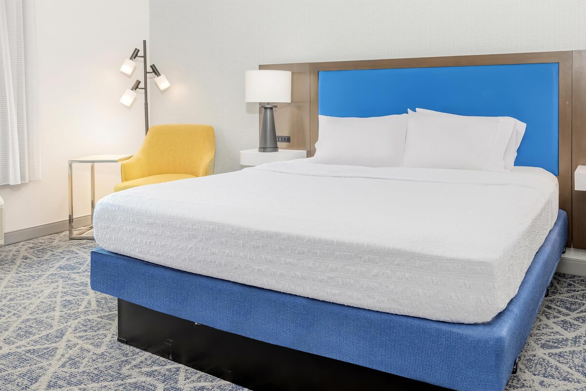 A hotel room with a large bed, a nightstand with a lamp, a chair, and a wall-mounted phone.