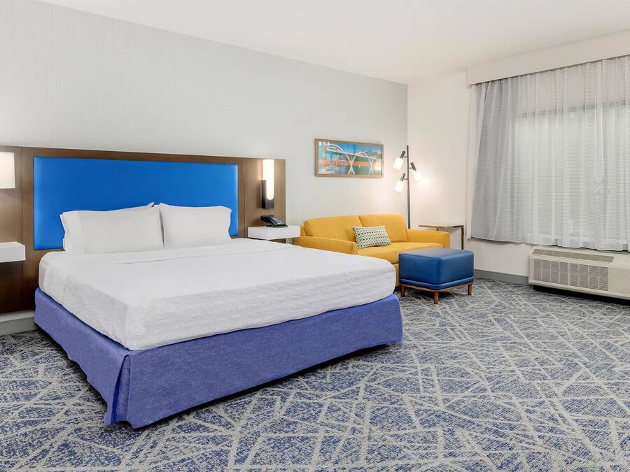 A hotel room with a bed, a bedside table, a lamp, a sofa, a chair, a radiator, and a window with curtains.