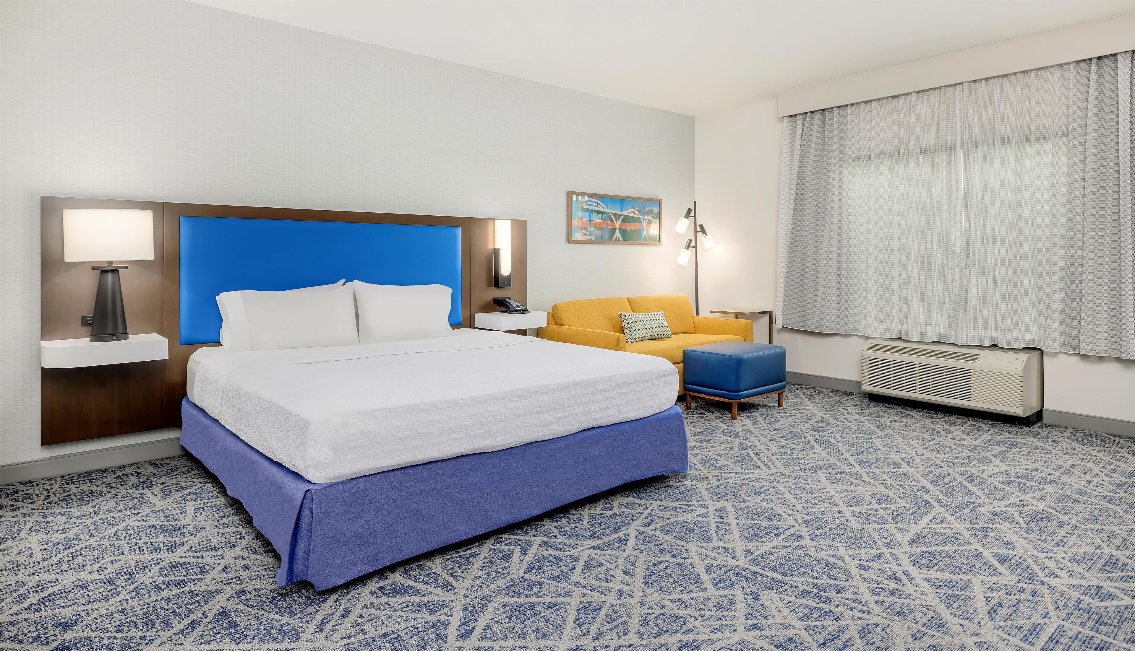 A hotel room with a large bed, a bedside table with a lamp, a small table with a chair, a sofa, and a window with curtains.
