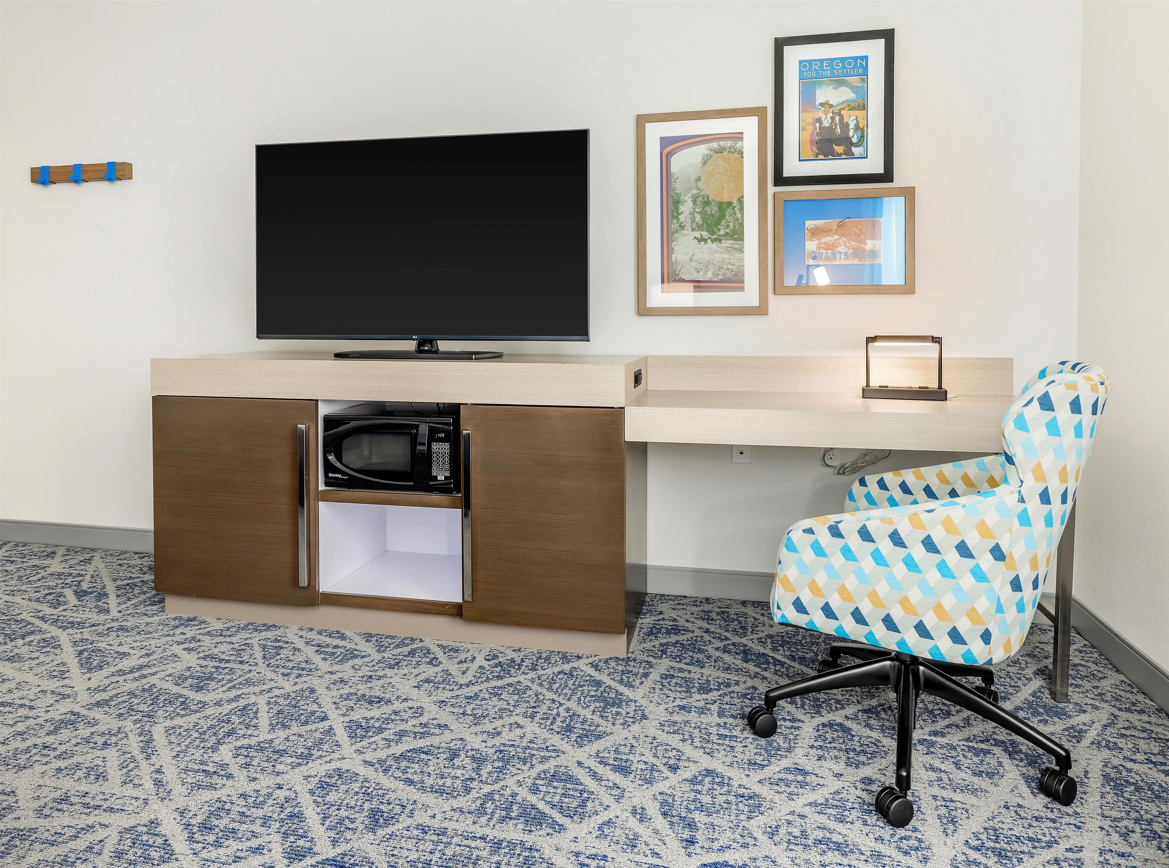 Une chambre d’hôtel moderne dotée d’un téléviseur à écran plat, d’un four à micro-ondes rangé dans un meuble, d’un tapis à motifs et d’une chaise de bureau colorée.