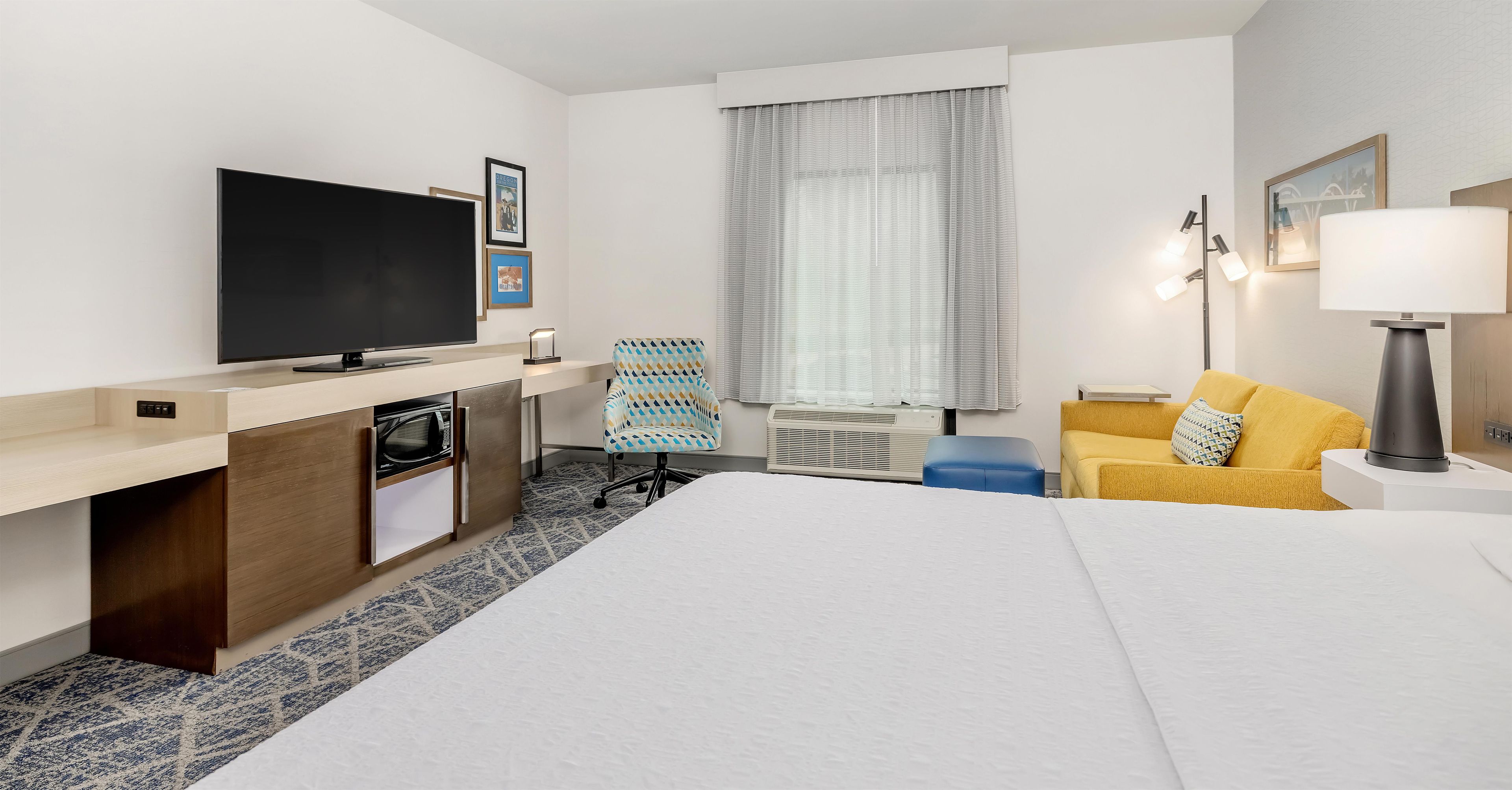 Habitación de hotel con cama, escritorio con televisor de pantalla plana, microondas, silla, sofá, lámpara y ventana con cortinas.