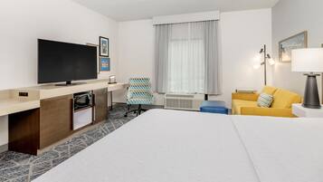 Habitación de hotel con cama, escritorio con televisor de pantalla plana, microondas, silla, sofá, lámpara y ventana con cortinas.