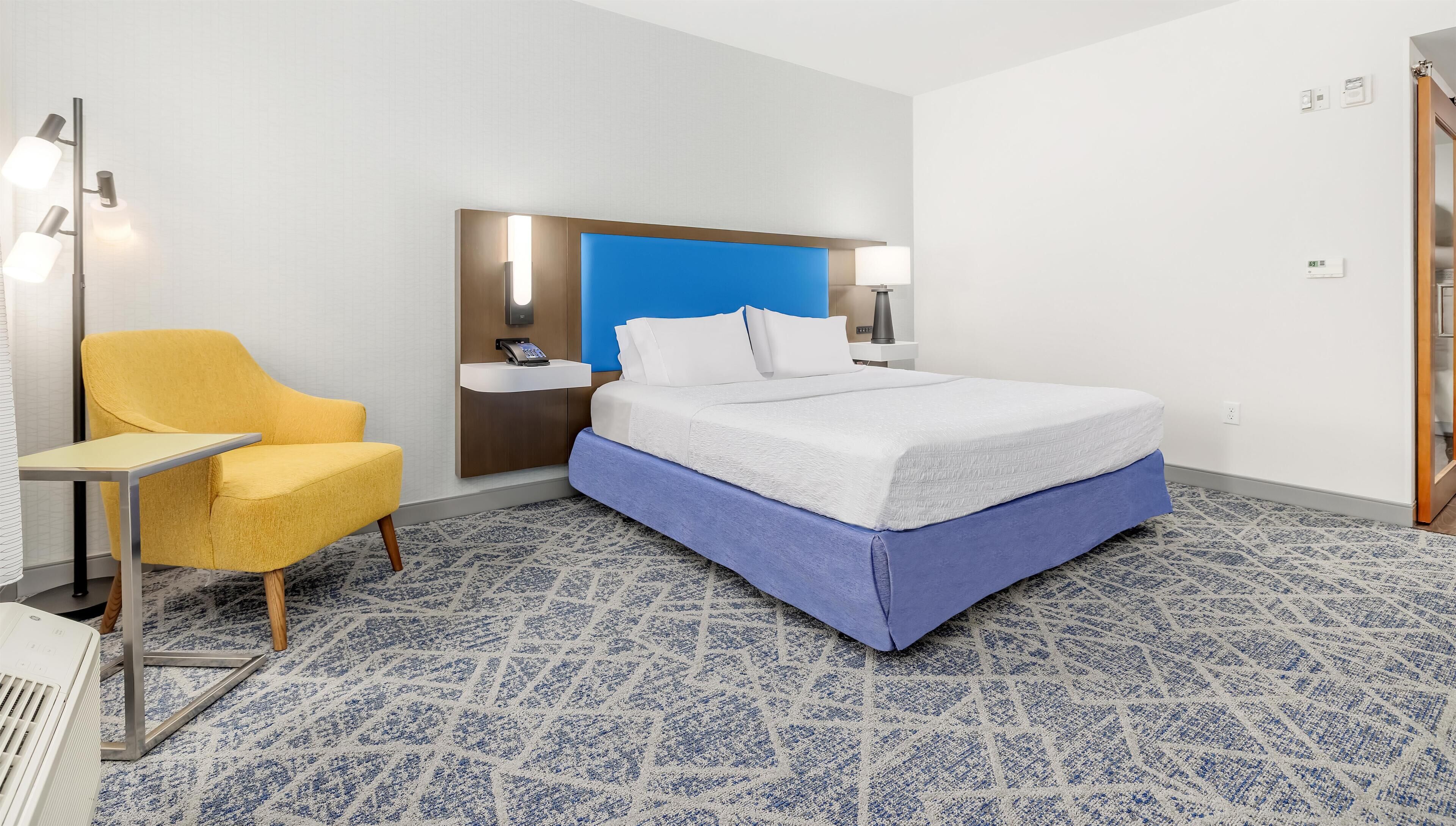 A hotel room with a bed, a desk, a chair, a bedside table, a wall-mounted phone, and a blue accent wall.