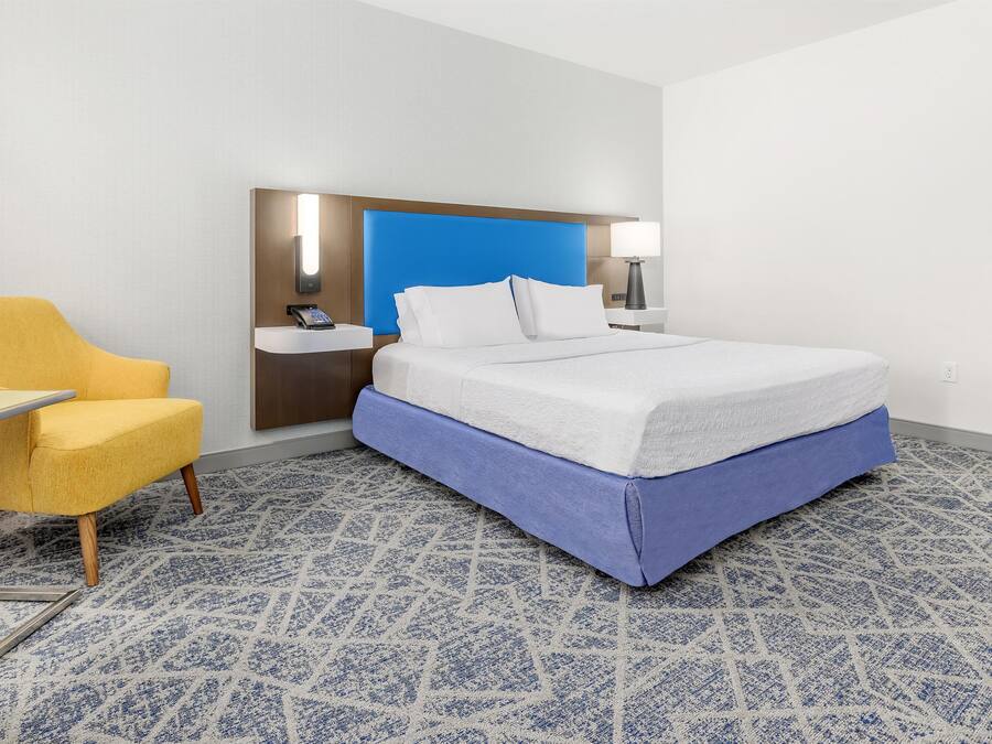 A hotel room with a bed, a desk, a chair, a bedside table, a wall-mounted phone, and a blue accent wall.