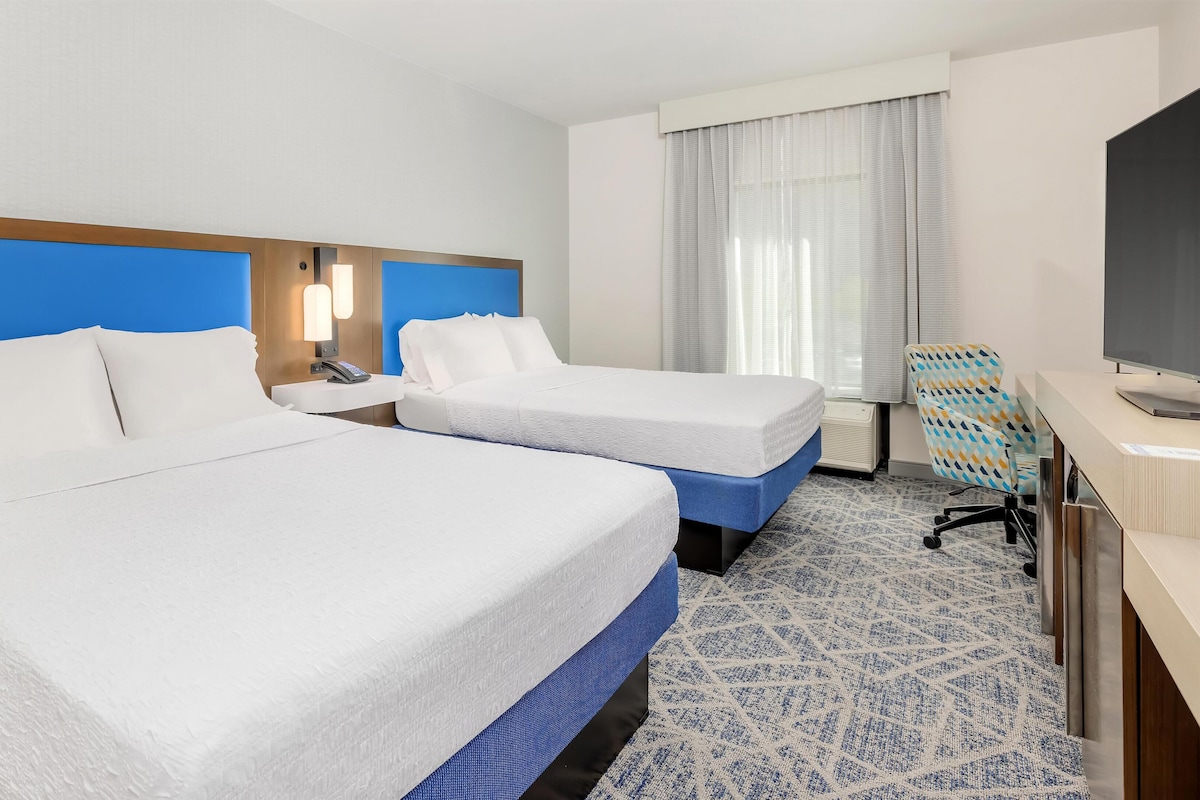 A hotel room with two beds, a desk, a chair, and a TV.