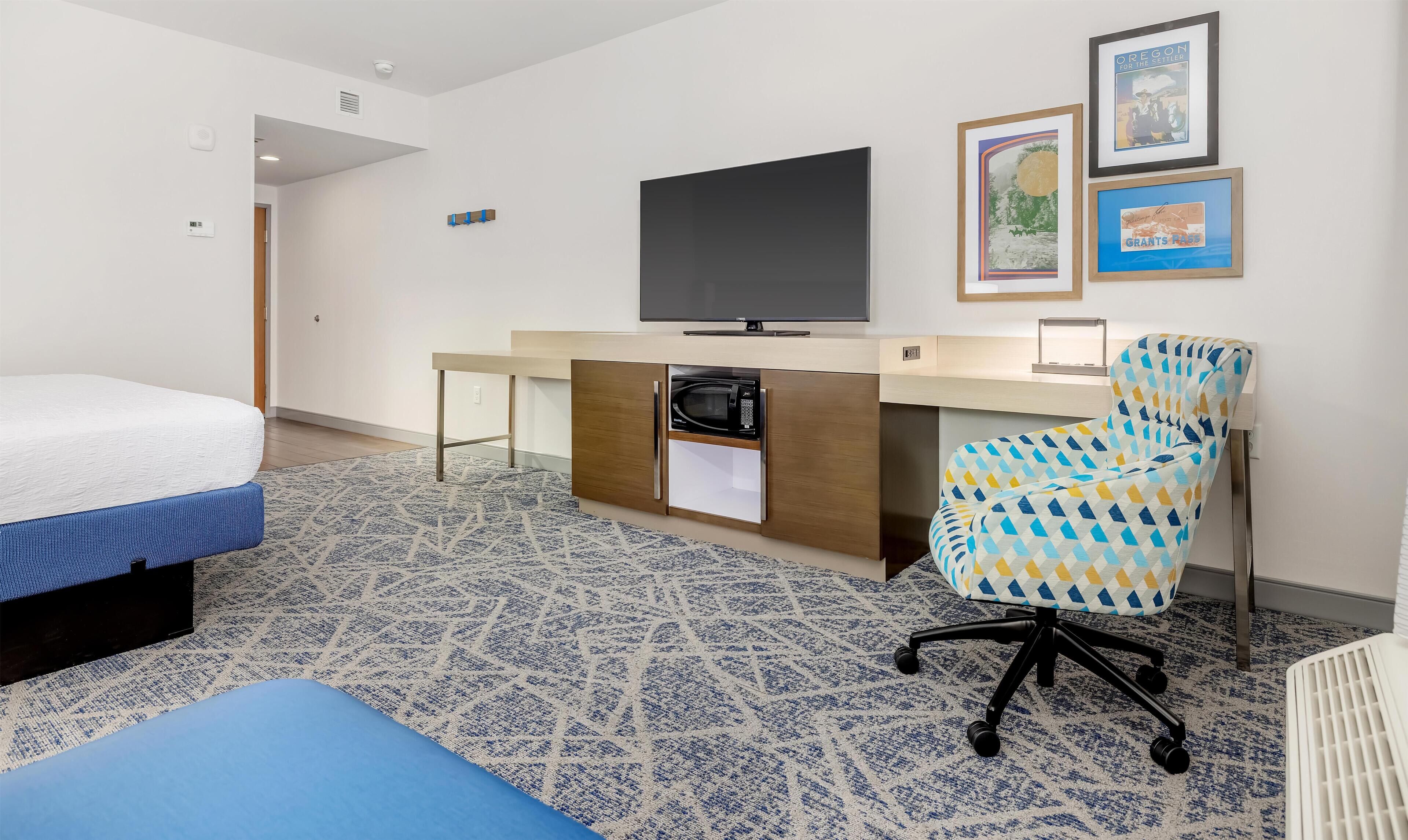 A hotel room with a bed, a desk with a TV, a microwave, and a chair.