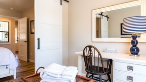 Une chambre d’hôtel avec un lit, une commode blanche à tiroirs, un miroir, une lampe, une chaise et un panier en osier contenant des serviettes.