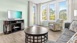 A living room with a TV, a round table, a sofa, and a chair. - Great Bay Views, Steps to Baytowne Wharf, Balcony, Beach Tram, Pool, Hot Tub. (Miramar Beach)