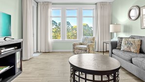 A living room with a sofa, armchair, round table, TV, and large windows with curtains. - Great Bay Views, Steps to Baytowne Wharf, Balcony, Beach Tram, Pool, Hot Tub. (Miramar Beach)
