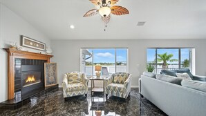 A living room with a fireplace, two armchairs, a sofa, and large windows offering a view of the outdoors. - Million dollar house on main river w/ million dollar view! Pool, Dock, Kayaks. (Crystal River)
