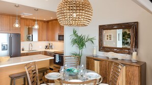 A modern kitchen with wooden cabinets, a central island, and a dining area with a glass table and chairs. - Beachfront Condo B303 - December 13-27 Still Available! (Kihei)